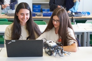Schulinformationen, zwei Schlerinnen mit PC und Roboter